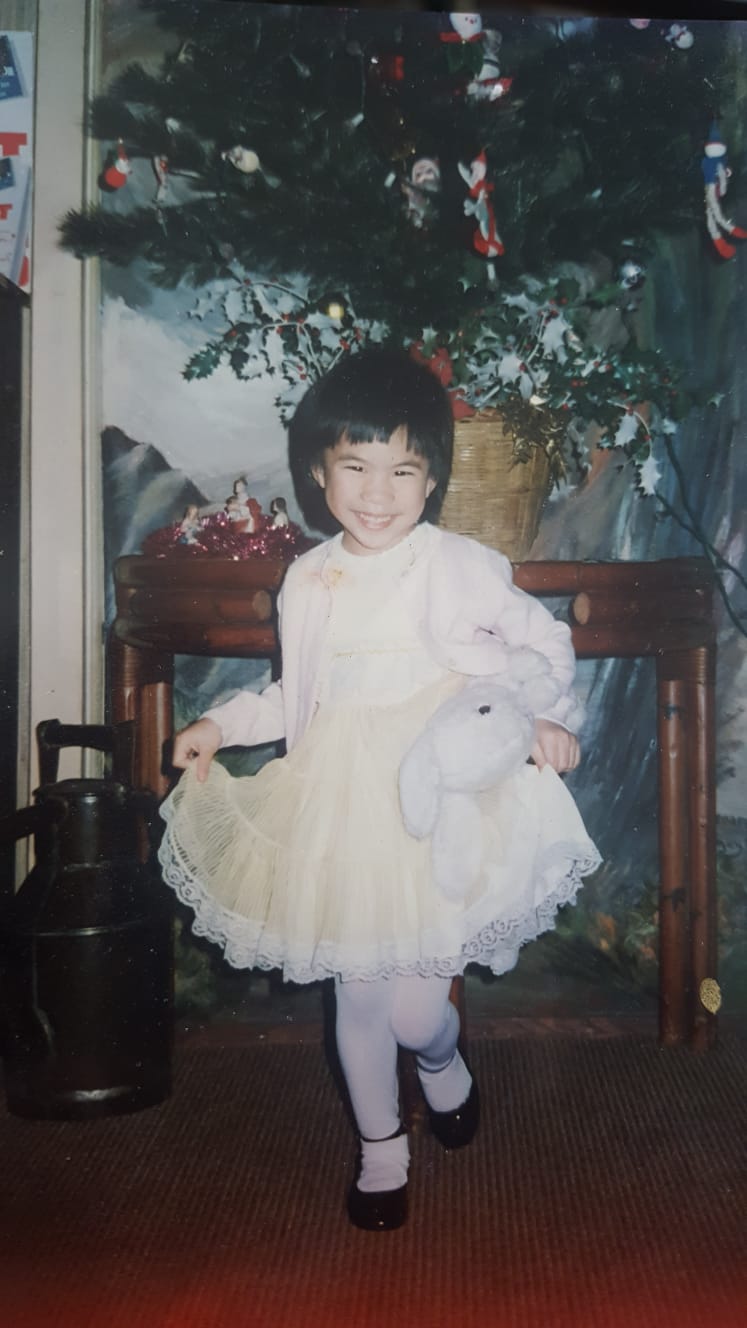 Grace enfant, souriante, en robe blanche à volants et une peluche sous le bras, devant un sapin de Noël